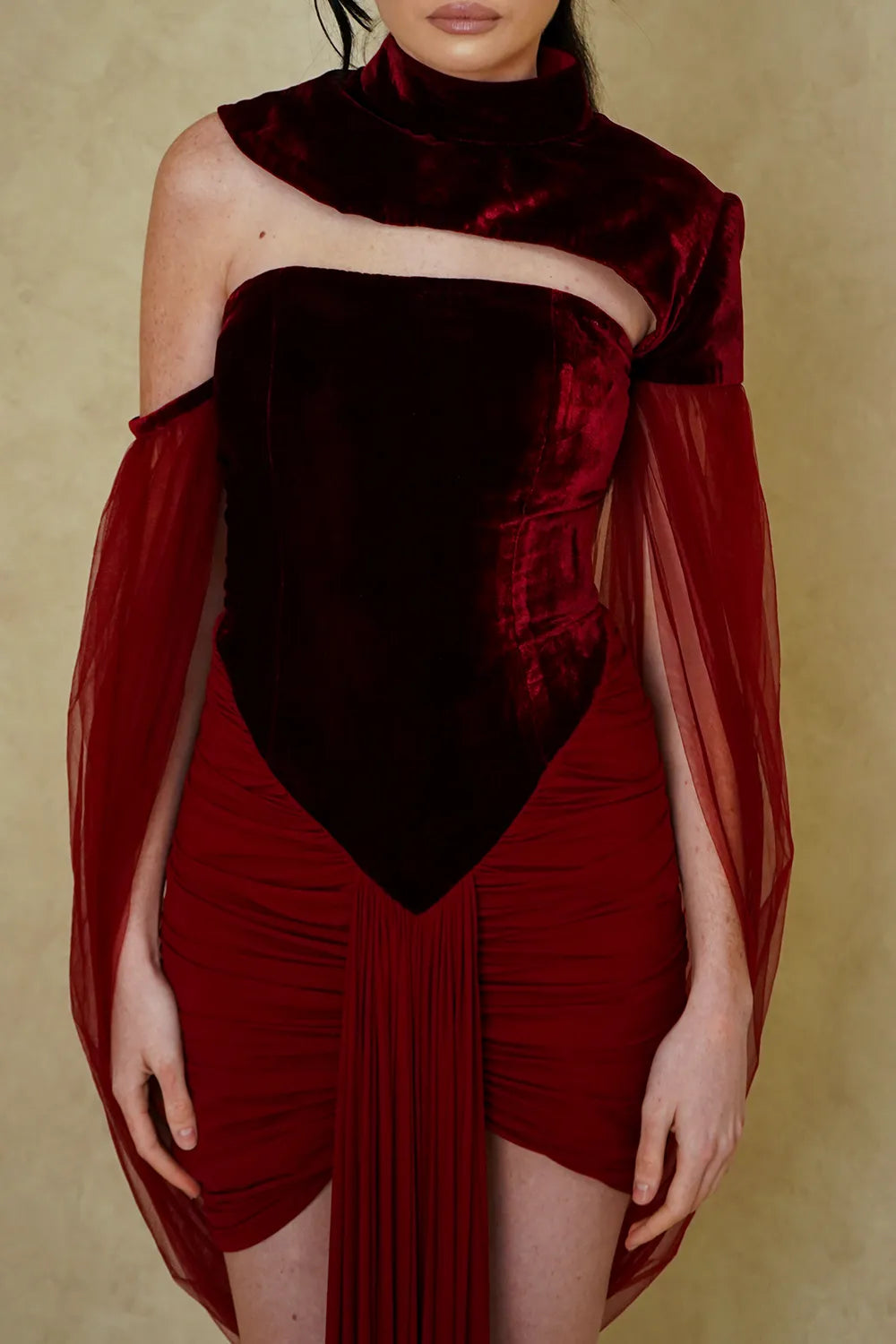 A model wearing a deep burgundy velvet off-the-shoulder mini dress with sheer tulle cape sleeves, set against a textured beige background.
